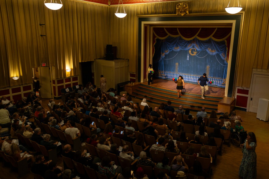 The house lights reveal a theater with a crowded audience as three people exit the stage with buckets to collect votes from spectators.