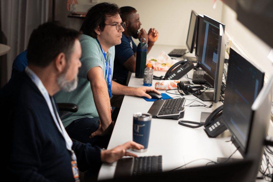 Three men work at computers along a desk with monitors, keyboards, phones, and drinks in a dim office.