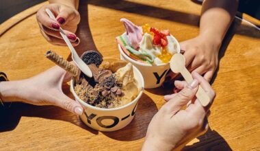 Two hands holding wooden spoons over Yo-Chi frozen yogurt cups topped with Oreo cookies, chocolate chips, wafer rolls, fresh strawberries, sour candy strips, and popping boba.