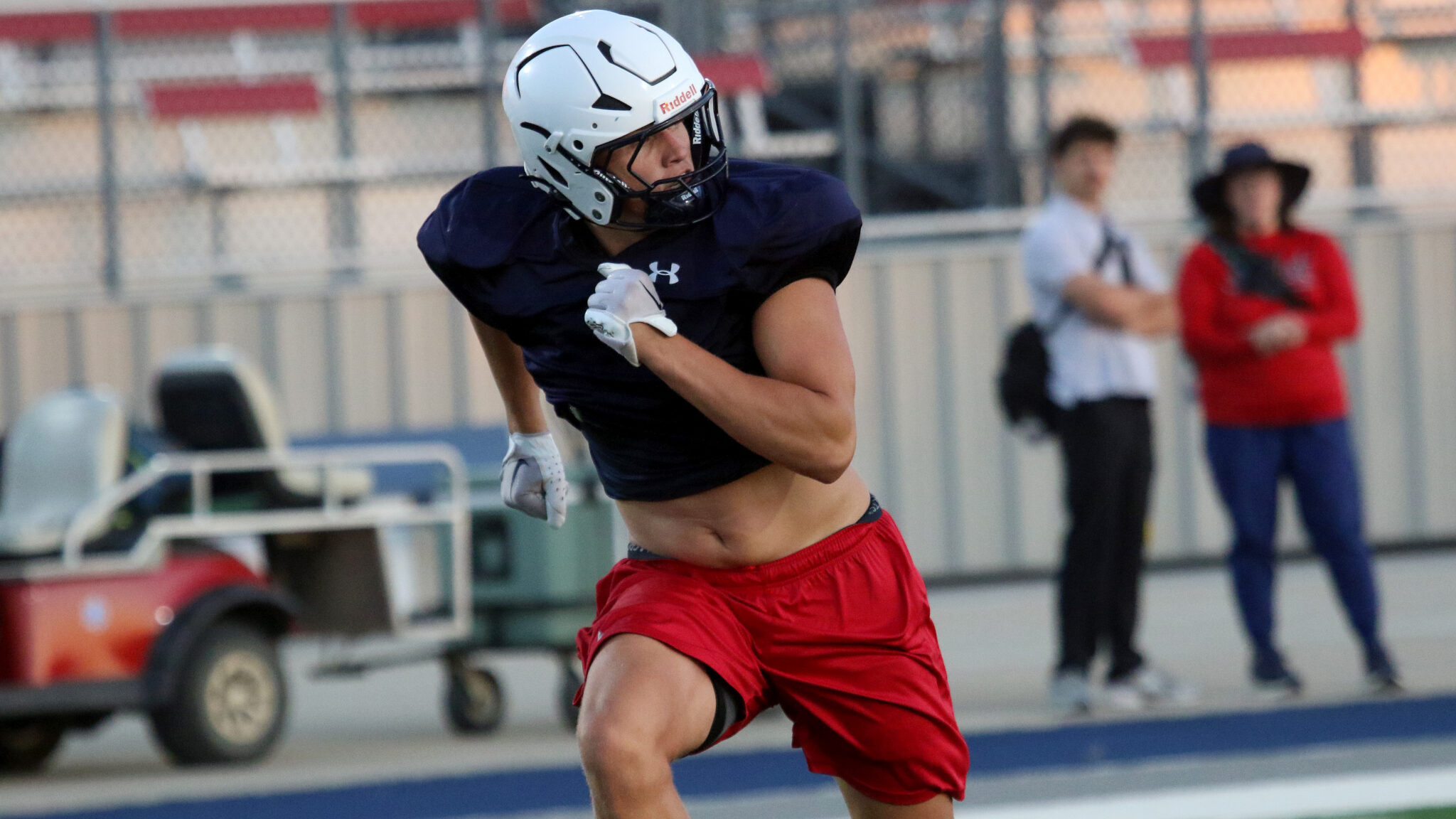 American football player in navy jersey and red shorts sprints on the field wearing a white helmet and gloves.