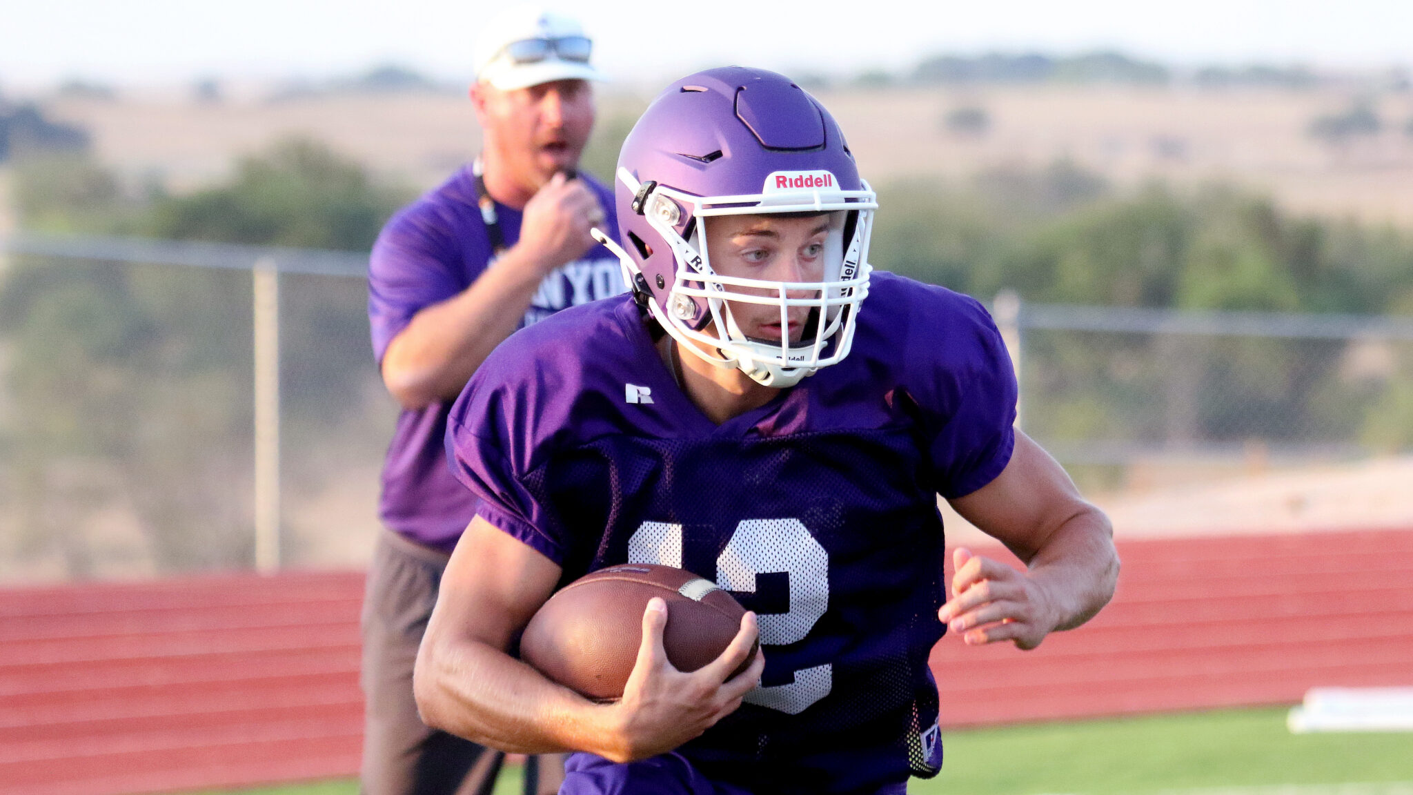 Football player in a purple jersey carrying a football while running on a track field, helmet on