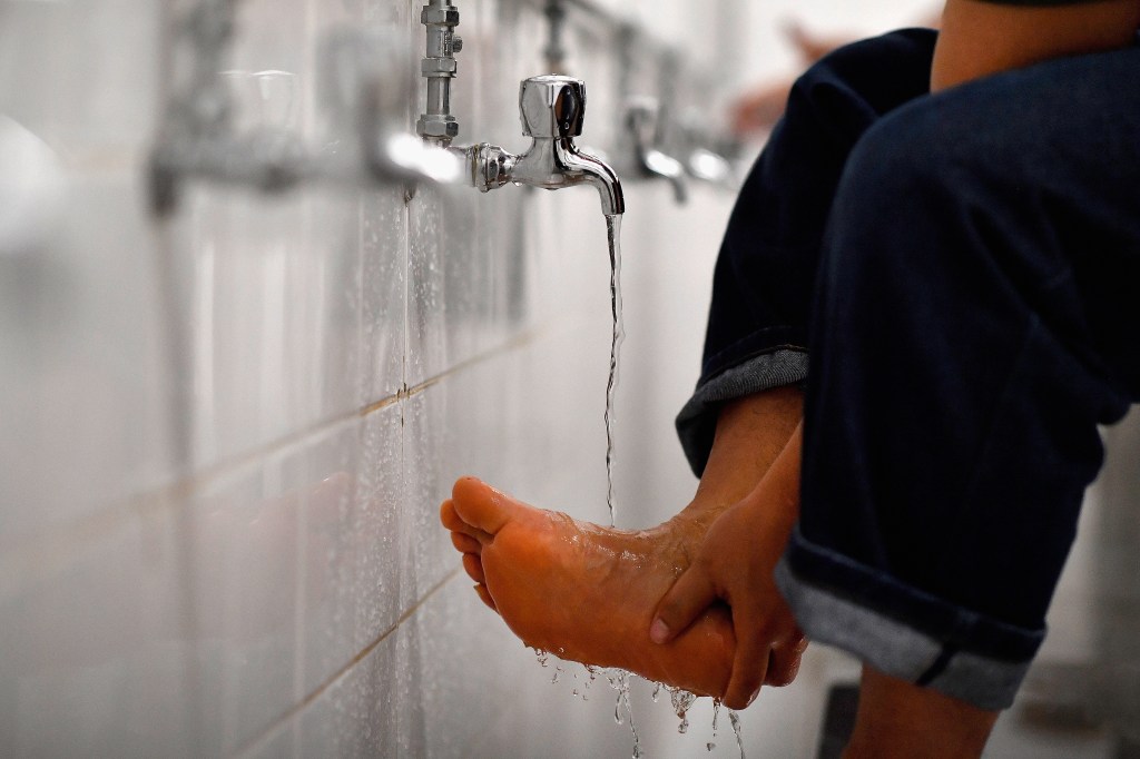 A person washing their foot with running water from a faucet in a room with white tiled walls.