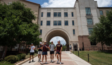 A group of students walk on a pathway toward a large Texas campus building with an archway, surrounded by trees and a partly cloudy sky.