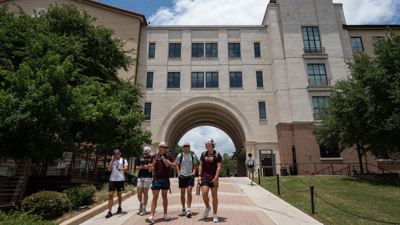 A group of students walk on a pathway toward a large Texas campus building with an archway, surrounded by trees and a partly cloudy sky.