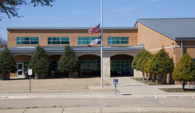 A brick school building with two flags at half-staff in front, surrounded by trimmed trees and a sidewalk under a clear blue sky.