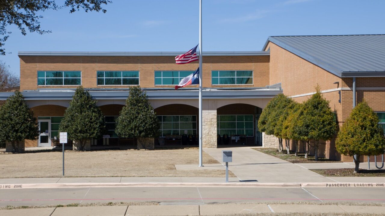 A brick school building with two flags at half-staff in front, surrounded by trimmed trees and a sidewalk under a clear blue sky.