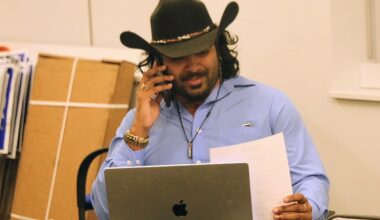 Man in a cowboy hat on a phone call at a desk, with a MacBook and papers in hand.
