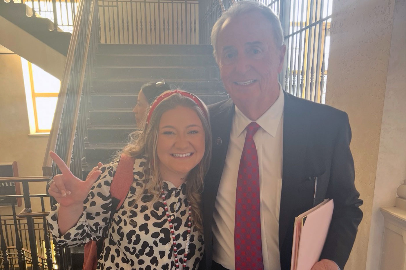 A student wearing a red backpack and a man in a suit smile together inside a stairwell area on campus, with the student making a “Guns Up” hand sign.