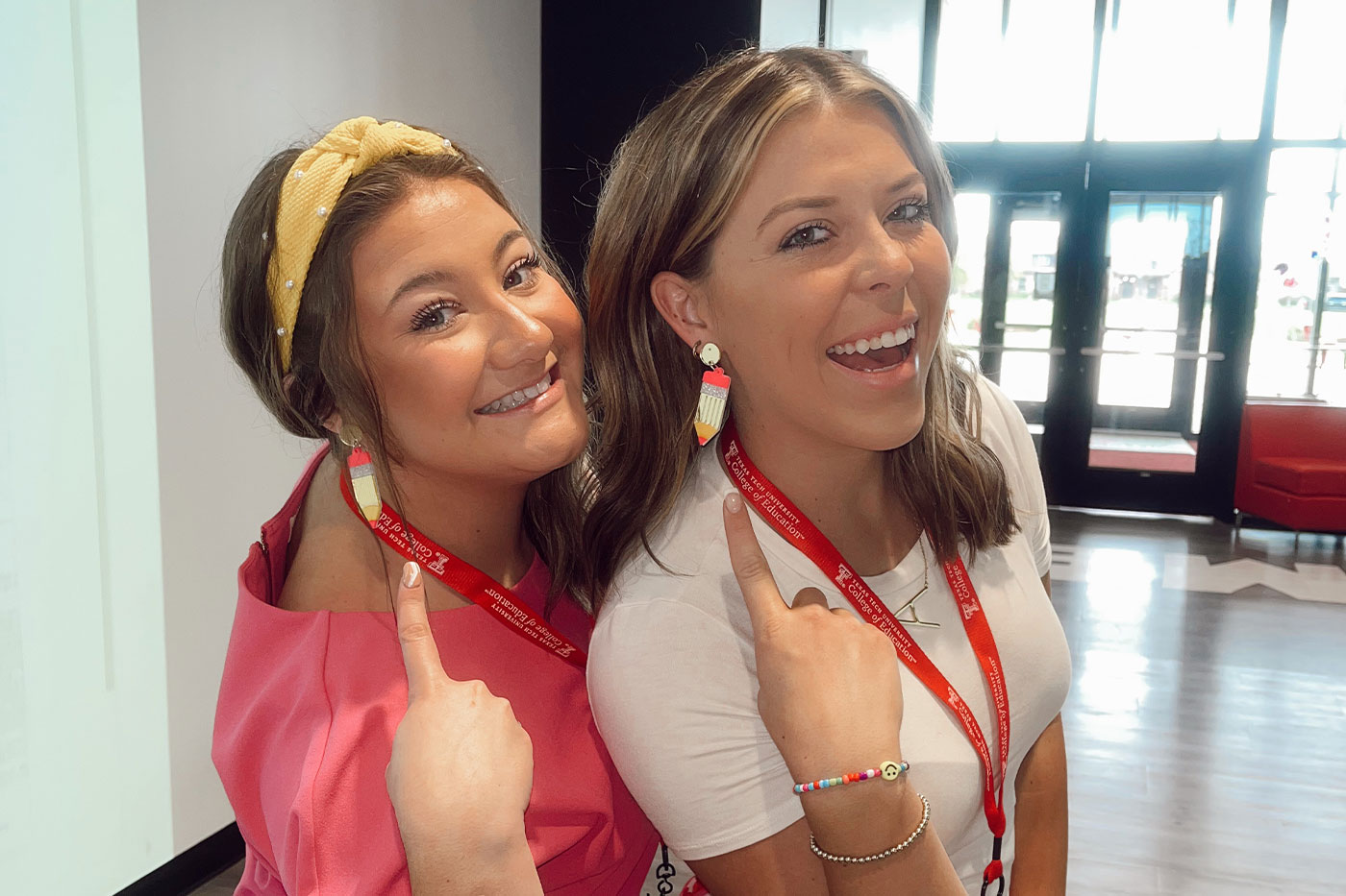 Two women wearing red Texas Tech lanyards smile and point to pencil-shaped earrings indoors.