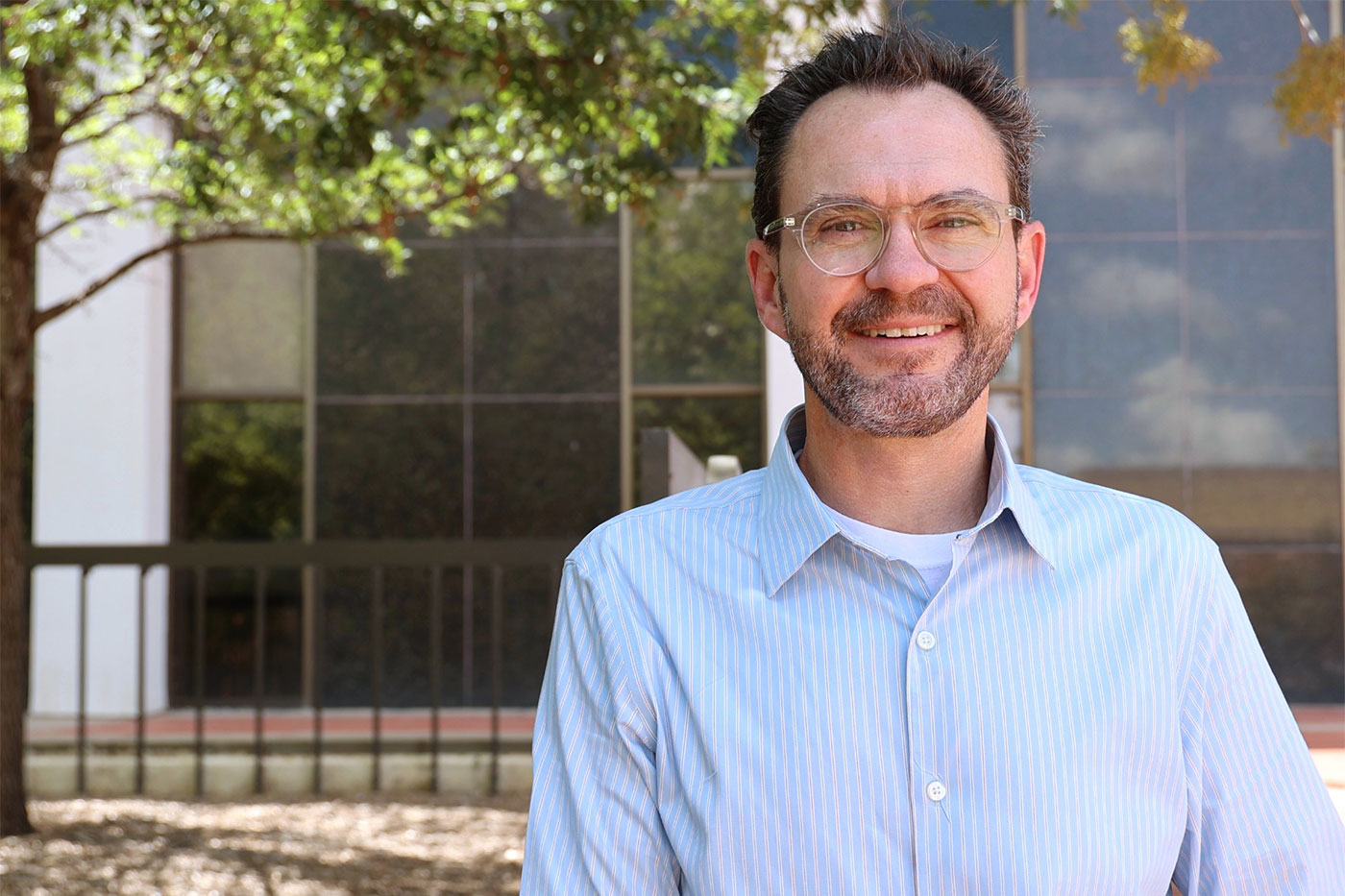 A man wearing glasses and a light blue striped shirt stands outdoors in front of a campus building, smiling toward the camera.