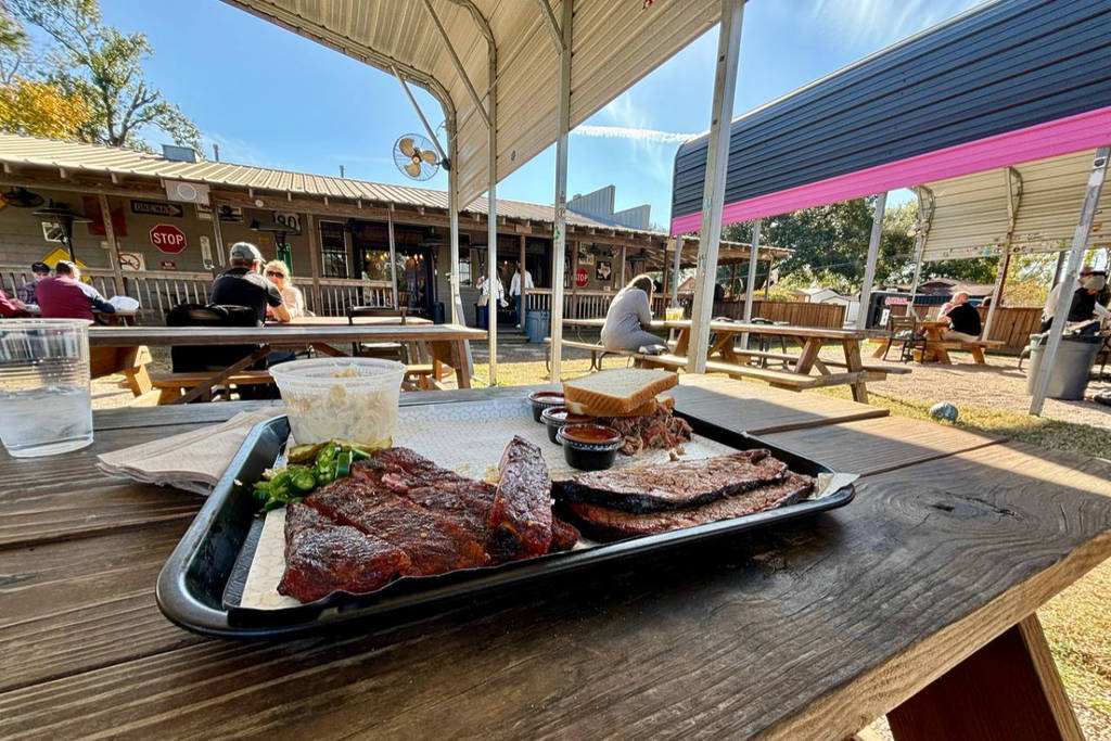 Image shows a plate of barbecue on a picnic table among a bag patio at Corkscrew BBQ in Houston