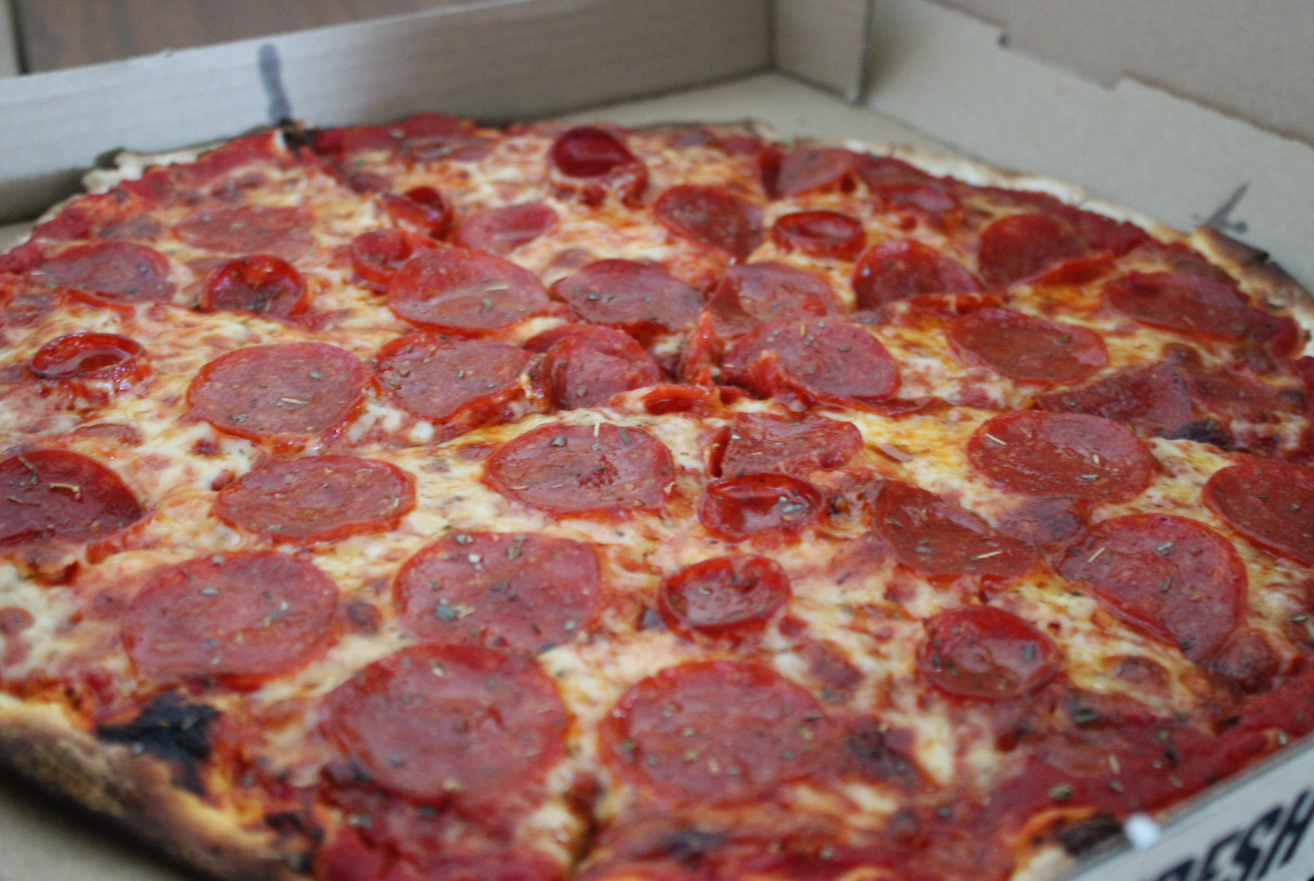 Ghost Pizza with pepporoni and hot cups laid across the top. Red sauce peeps above blackened thick crust.
