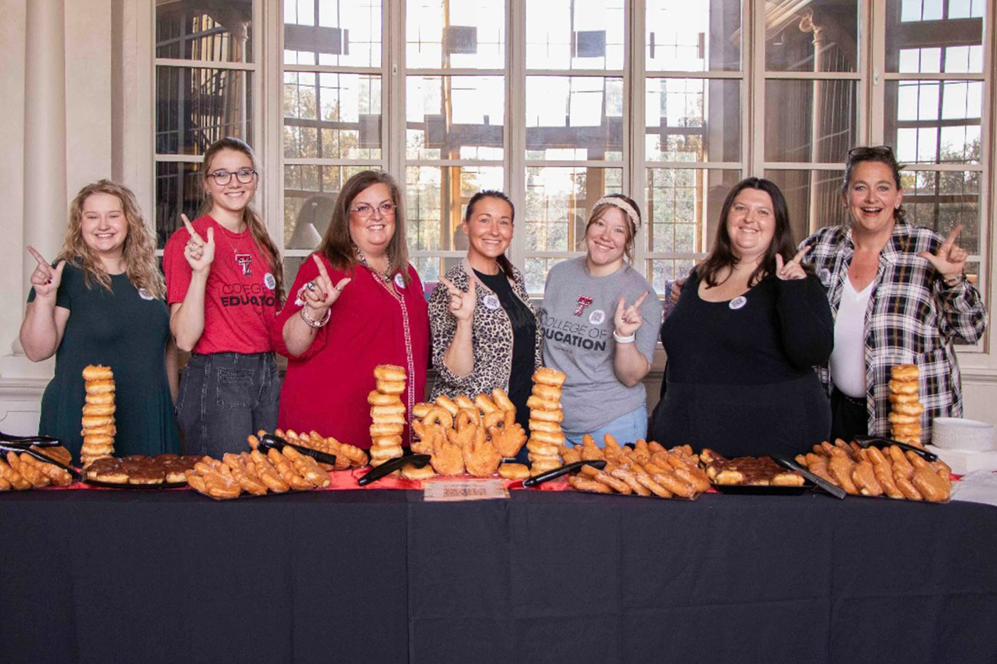 Seven people stand behind a table filled with pastries, smiling and making “Guns Up” hand signs.