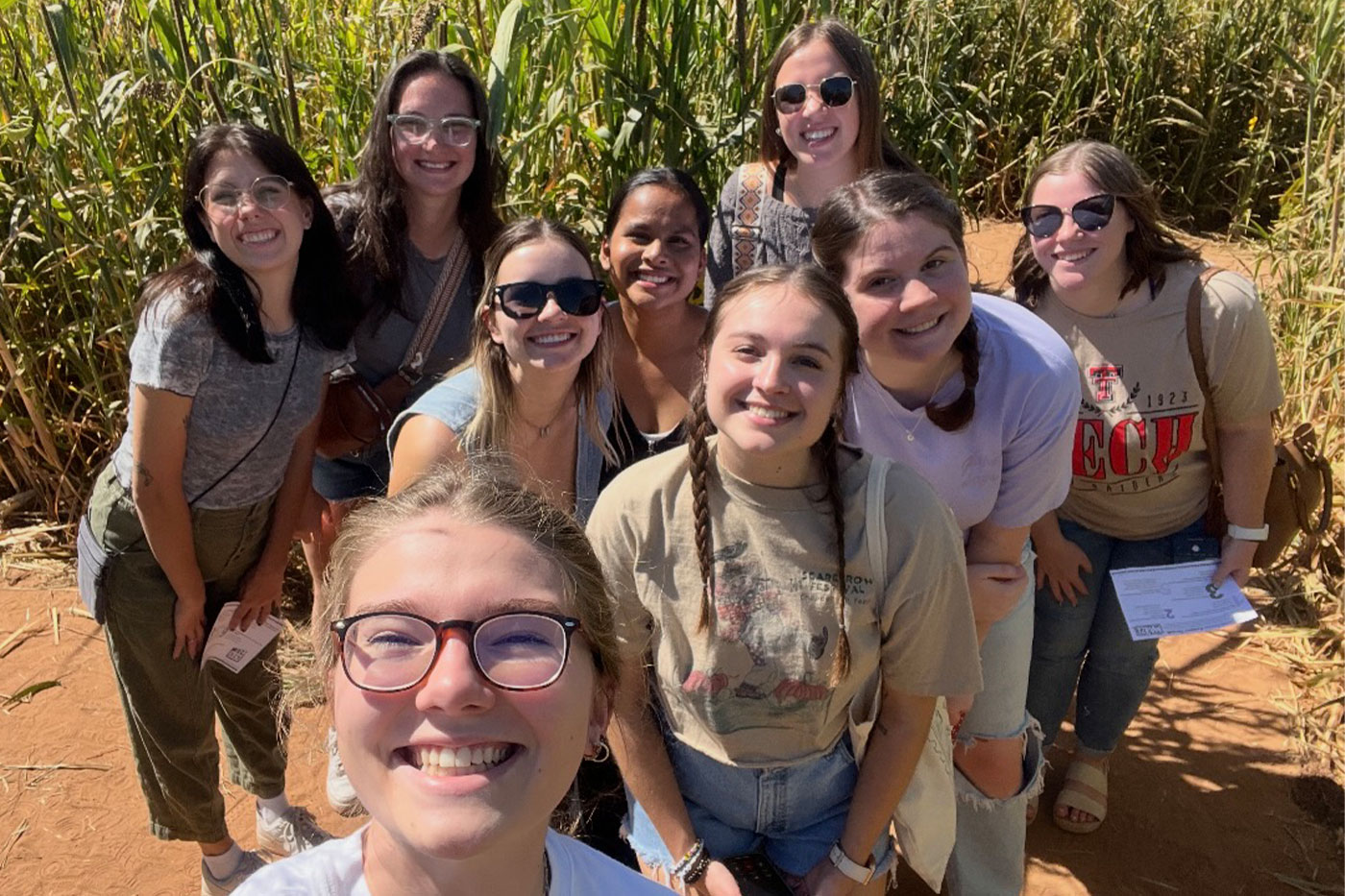 Nine students pose together for a selfie in a corn field on a sunny day, smiling toward the camera.