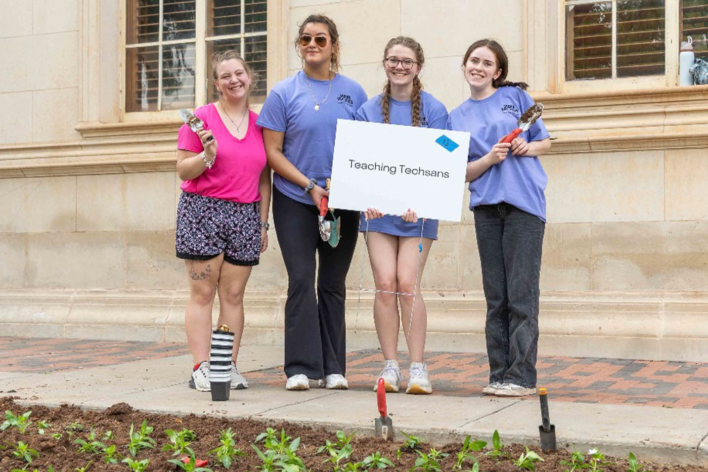 Four students pose beside a freshly planted garden bed outside a campus building, holding small gardening tools and a “Teaching Techsans” sign.