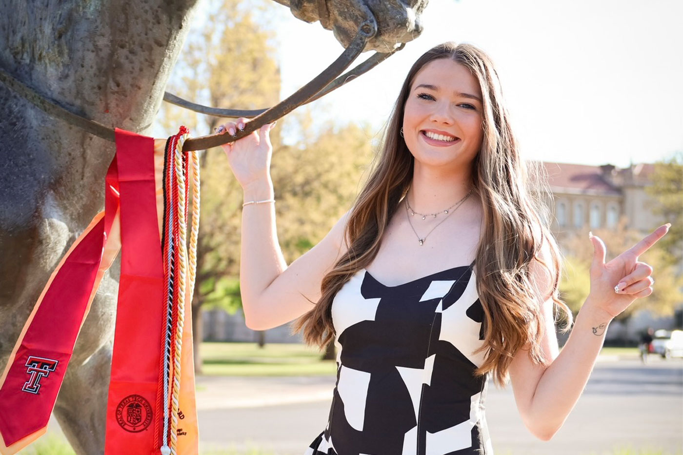 A Texas Tech graduate stands next to the Will Rogers statue, holding red and gold graduation stoles and cords while smiling and making a “Guns Up” hand sign.