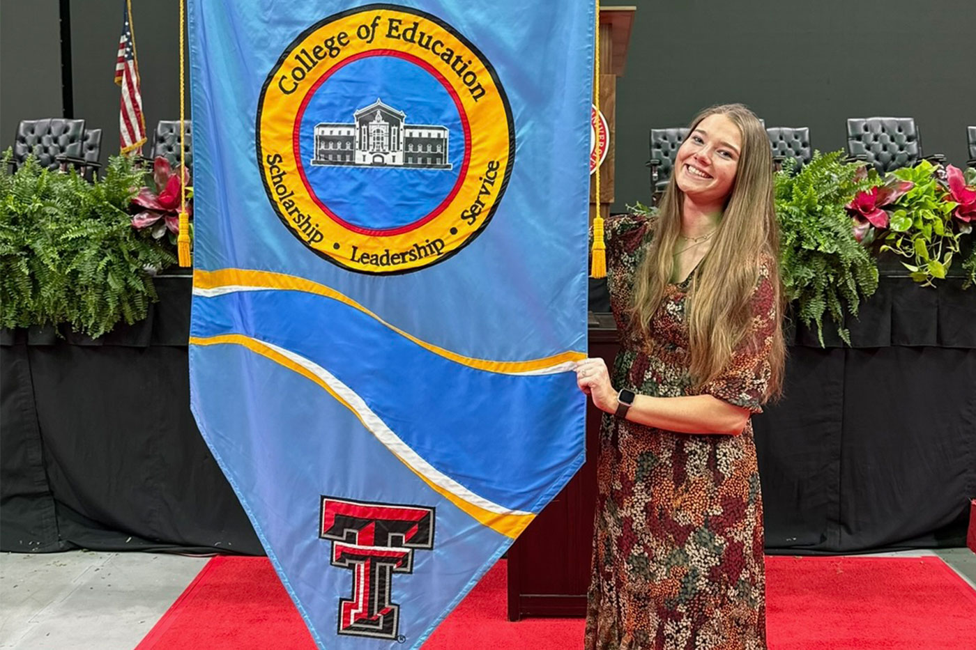 A student in a floral dress poses next to a blue Texas Tech College of Education banner that reads “Scholarship, Leadership, Service” during an indoor event.