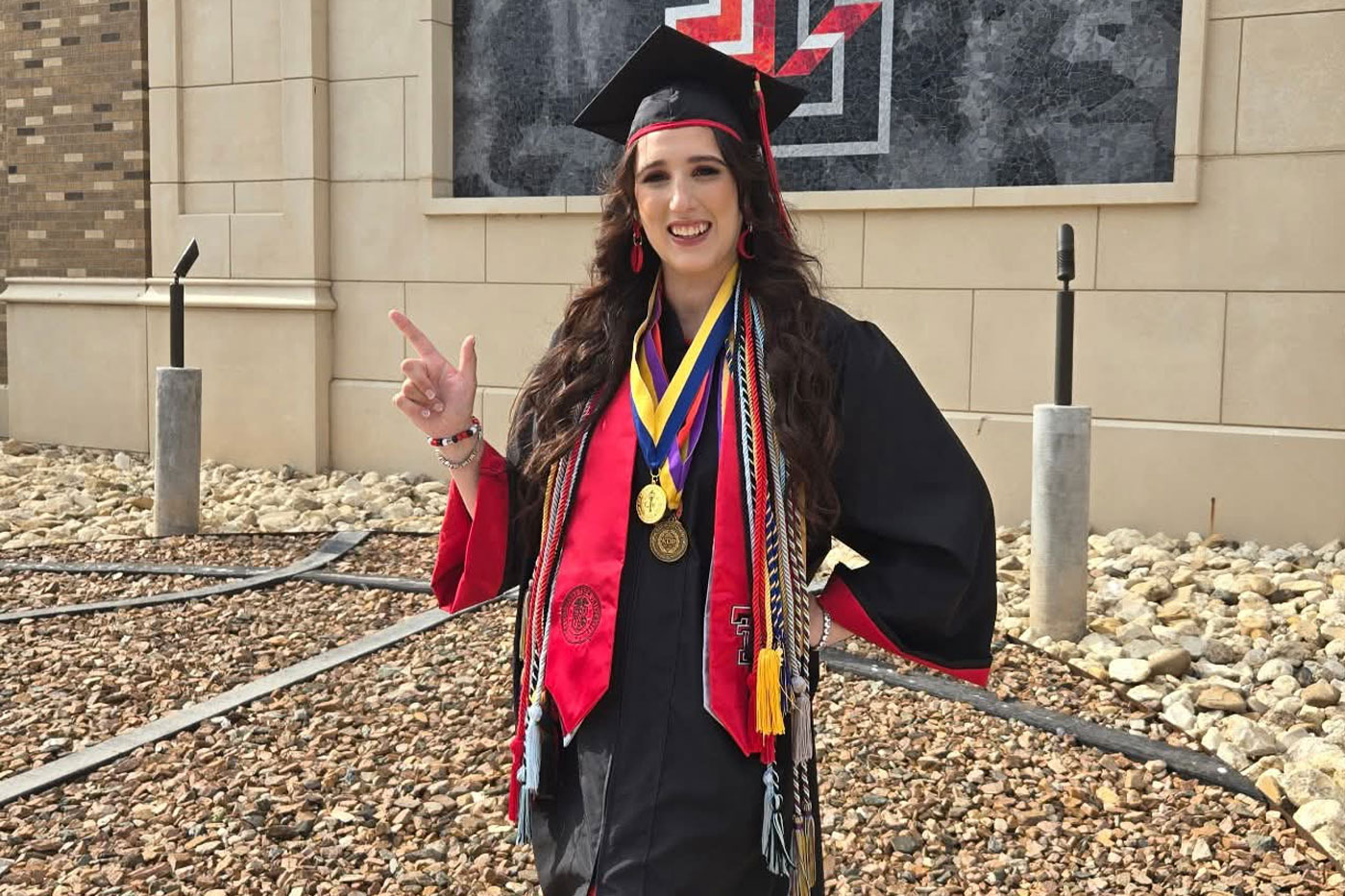 Texas Tech graduate, Katherine Kraatz, wearing a cap, gown, red stole, honors cords and medals stands outside a campus building and makes a “Guns Up” hand sign.