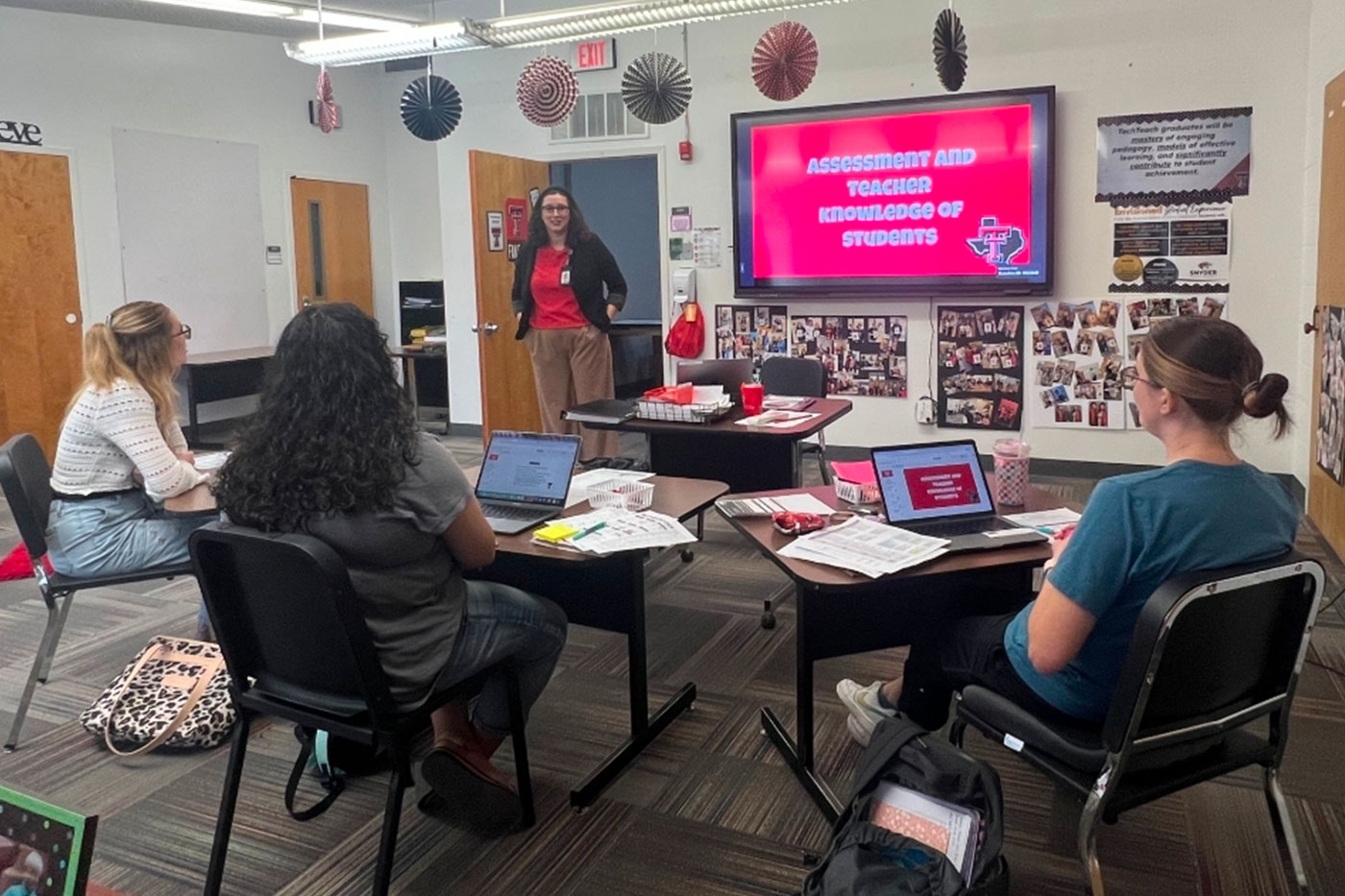 A presenter stands at the front of a classroom beside a screen that reads “Assessment and Teacher Knowledge of Students,” while students sit at tables with laptops and printed materials.