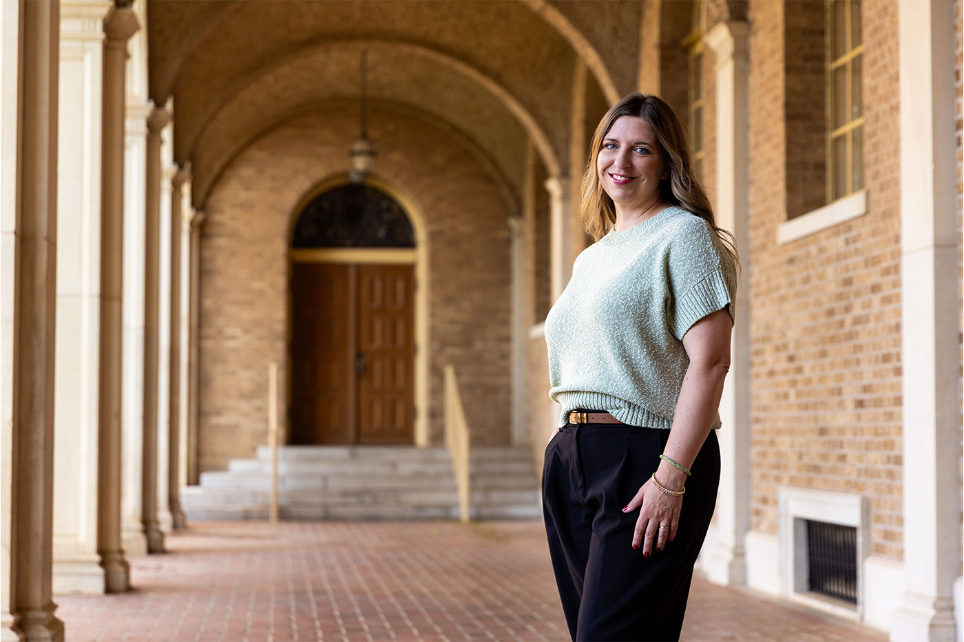 A woman in a light green sweater stands in a covered brick walkway with arched columns and a wooden doorway in the background.