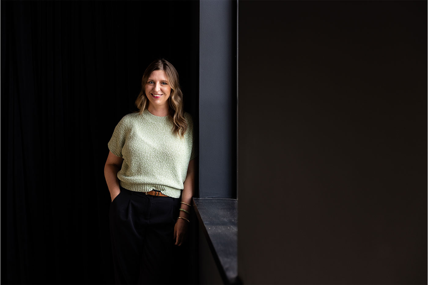 A woman in a light green sweater leans against a black wall.
