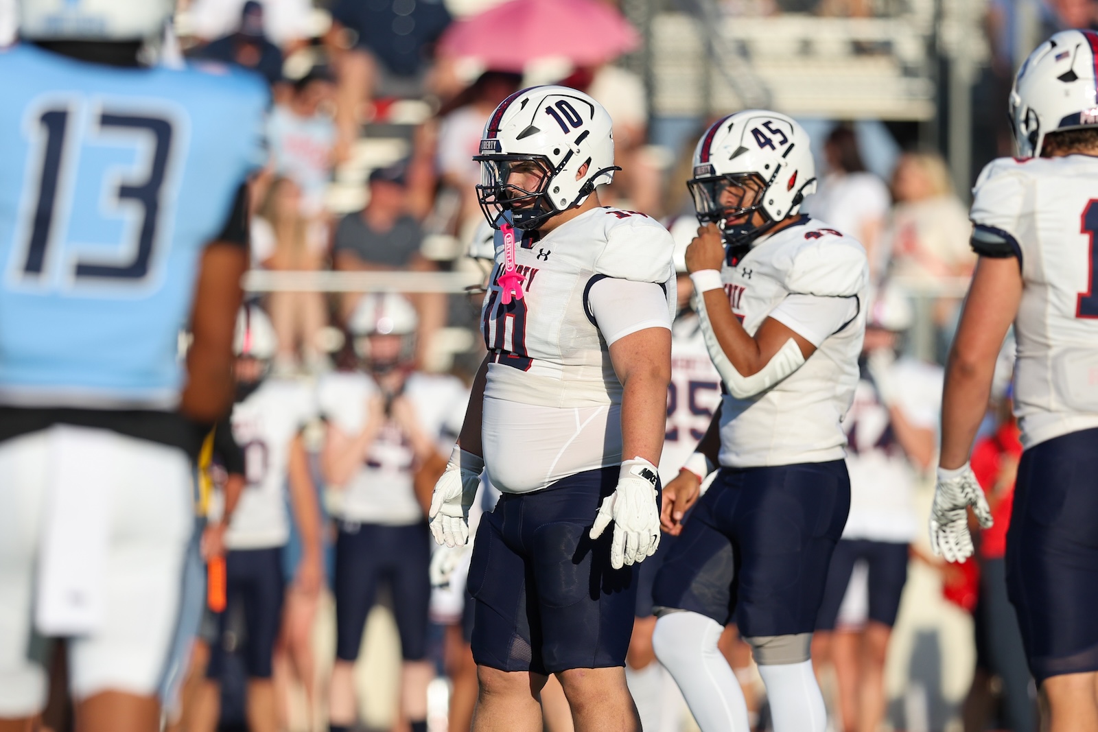 Football players in white uniforms and helmets standing on a sideline during a game, number 10 and 45 visible, crowd in background