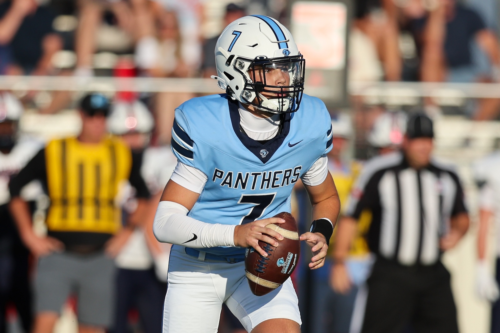 Panthers quarterback in a light-blue jersey with number 7 holds a football during a game on the field with a referee in the background.