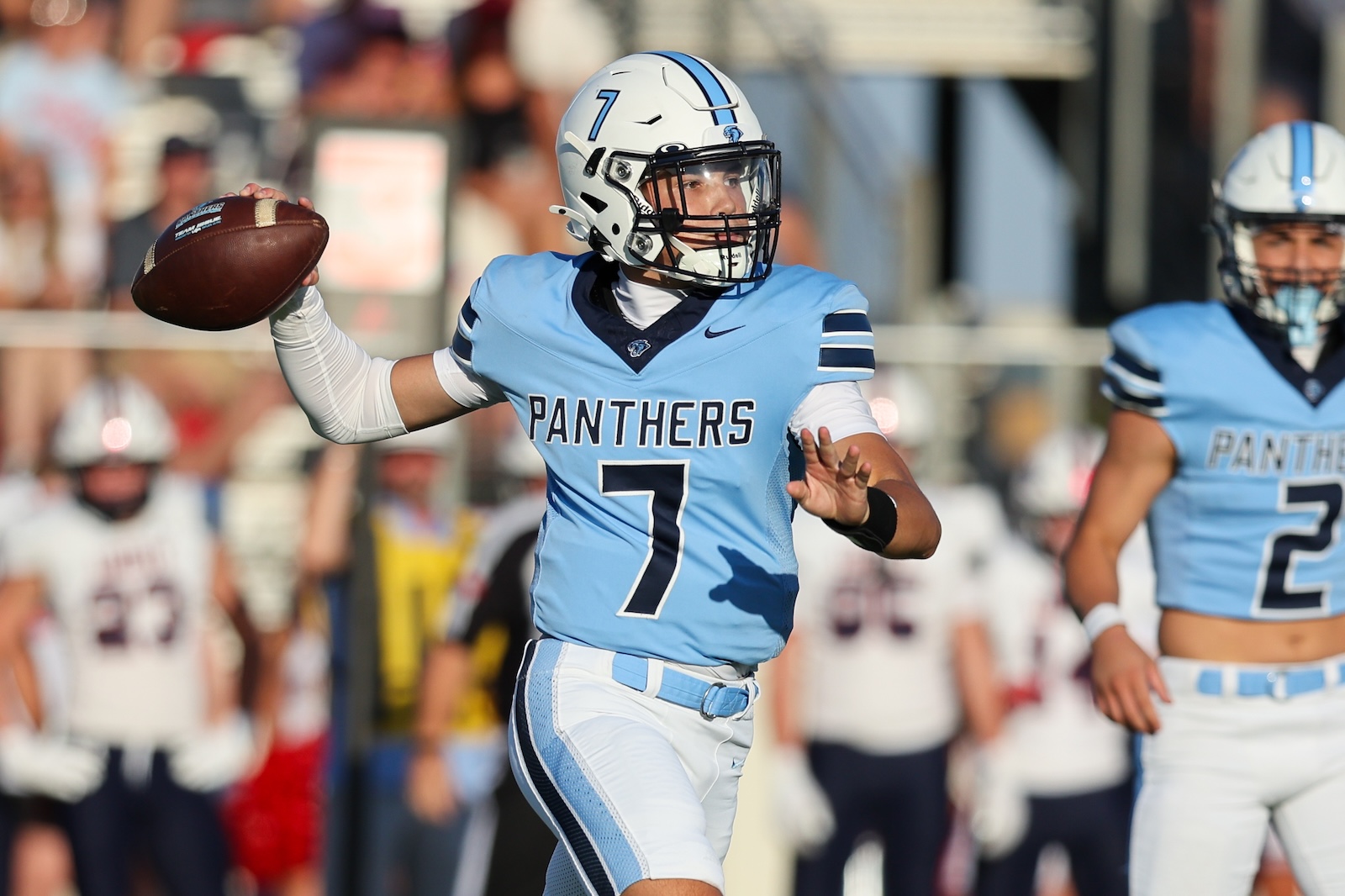 Panthers quarterback wearing number 7 in a light-blue uniform throws a pass during a game with a crowd in the background, and a teammate nearby.