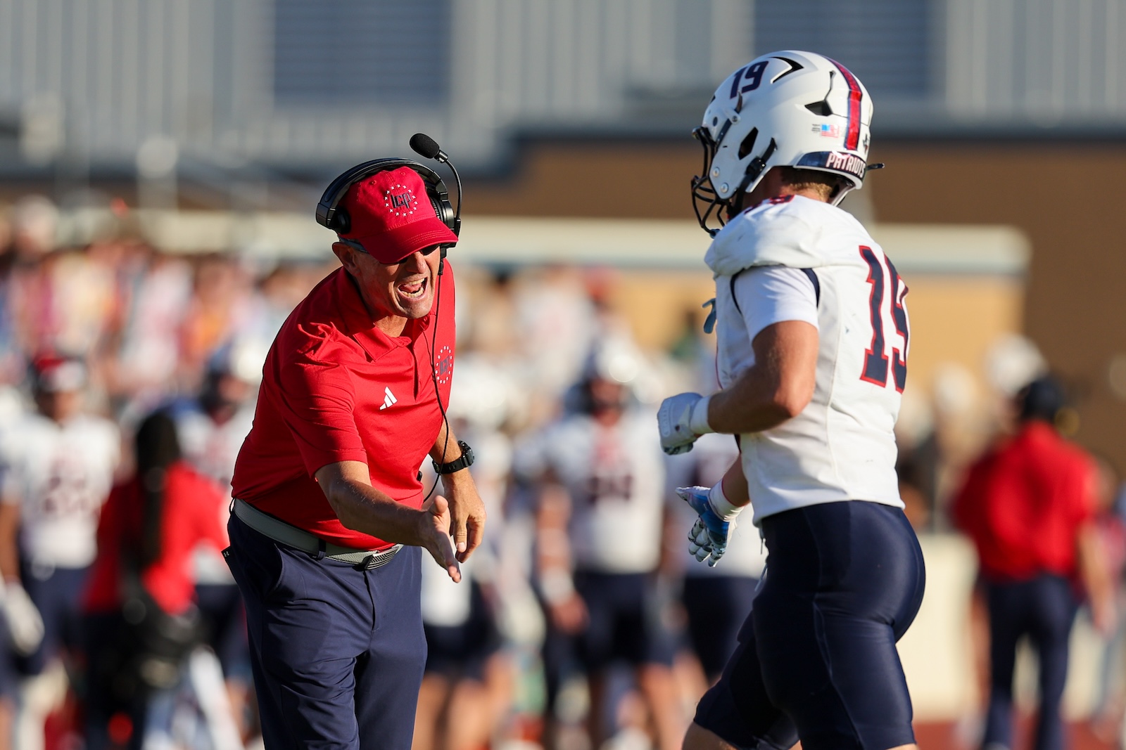 Football coach in red shirt and headset shouting at a player in white with a Patriot helmet during a game. Crowd blurred in the background.