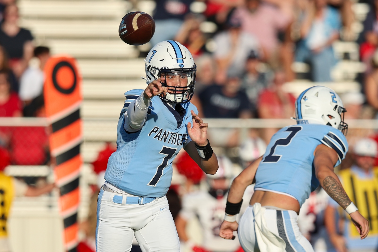 Quarterback in light blue Panthers uniform releasing a pass during a game, with teammates and a crowd in the background.