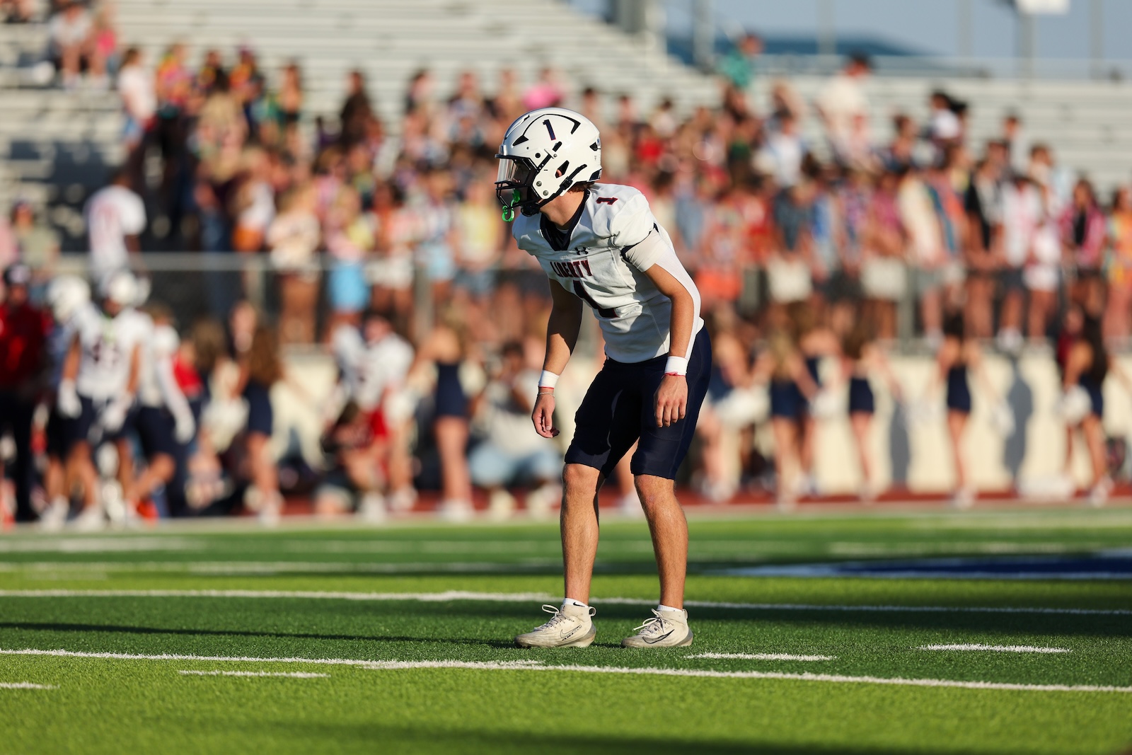 Football player in white jersey and helmet crouches on a green field with a crowded stadium in the background, ready for play.