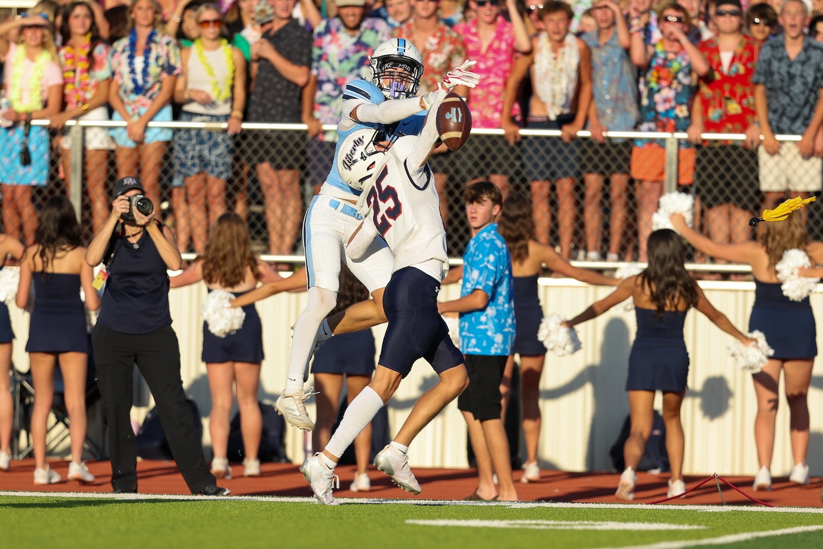 Two football players leap for a catches mid-air as a pass is contested, with a cheering crowd behind them on the stands.”