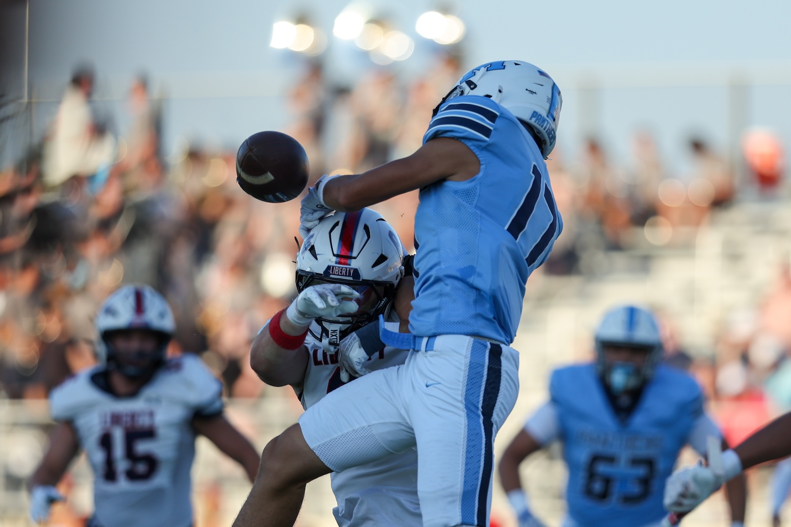 Two football players in light blue uniforms jump for a loose ball during a game, with a blurred crowd in the background.