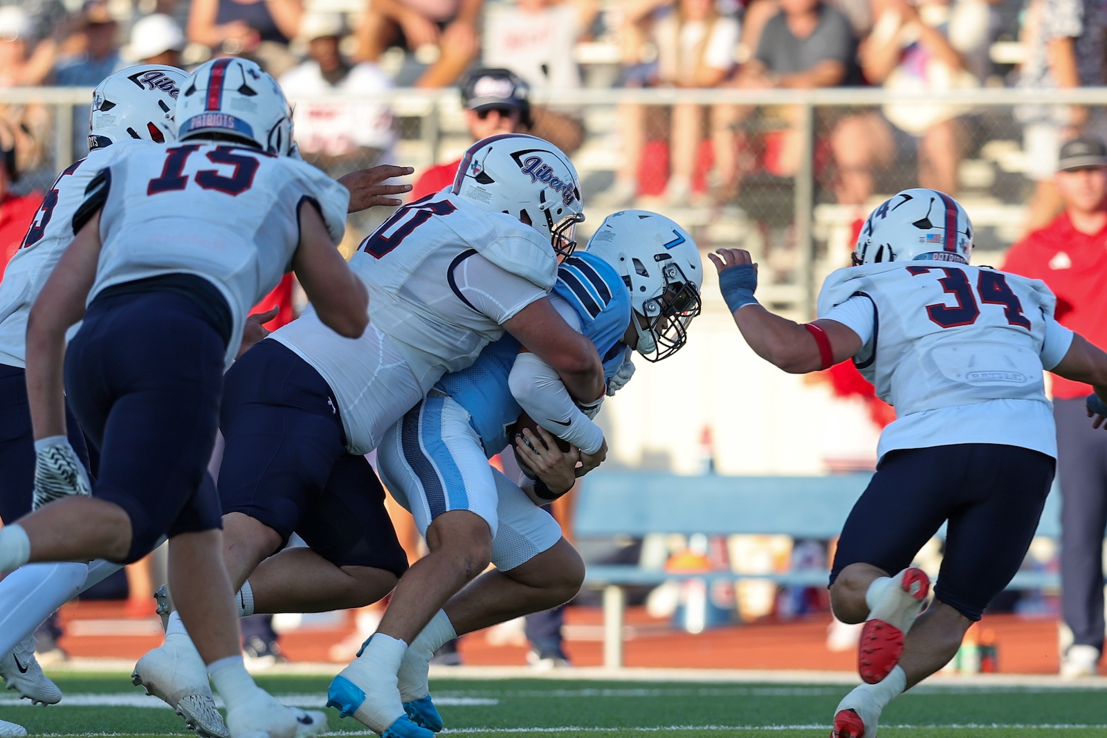 Football players in white and navy tackle a ball carrier in light blue during a game on a sunny day.