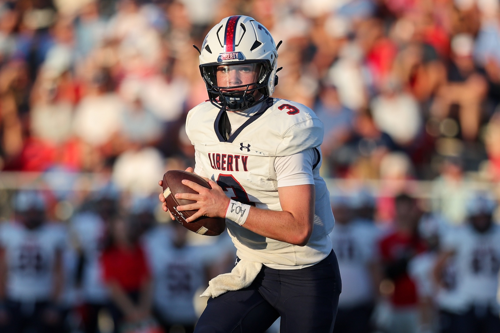 A Liberty football quarterback holding a football, running on the field with a crowd in the background.