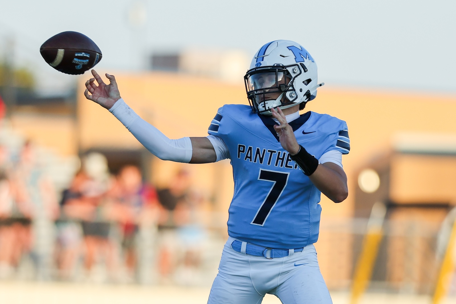 Quarterback in a light blue Panthers jersey (#7) throwing a football on a sunny field.