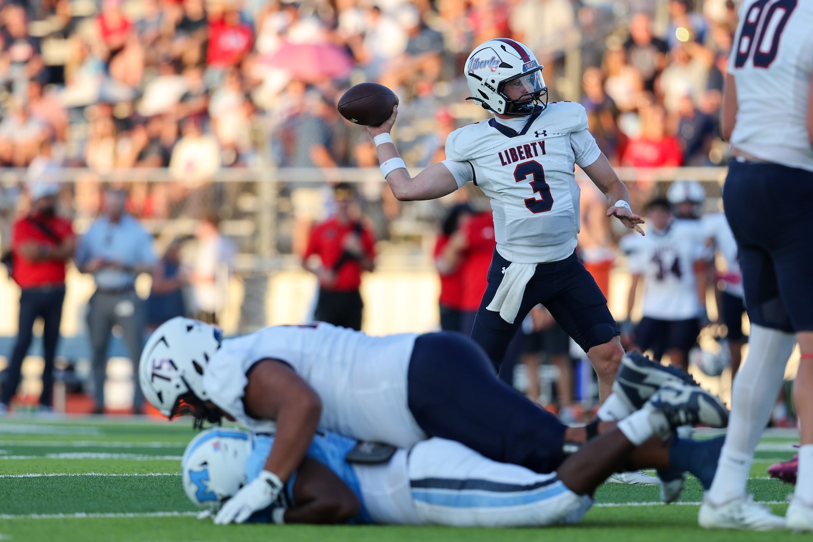 Quarterback in a Liberty white jersey (number 3) throwing a pass while teammates and opponents collide on the field, crowd in the stands behind.