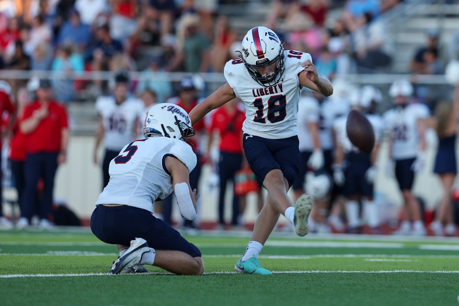Liberty football player in white #18 jumps to kick the ball while a defender crouches nearby on the field as the crowd watches in the stands.