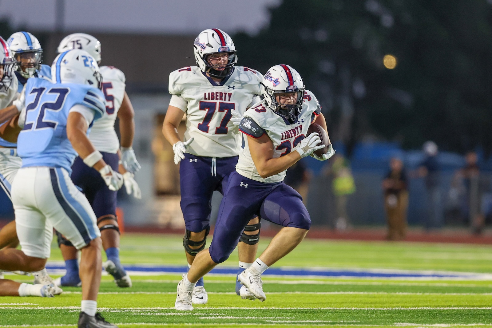 Liberty football player in white jersey carries the ball while teammates block on a bright green turf field at dusk/dawn gymnastics-style lighting.
