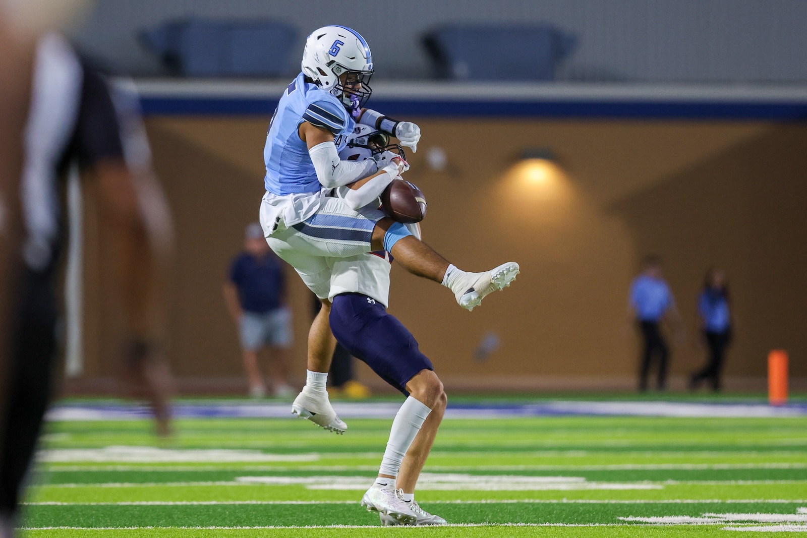 Two football players in blue and white jerseys contest a loose ball mid-air on a brightly lit indoor field.