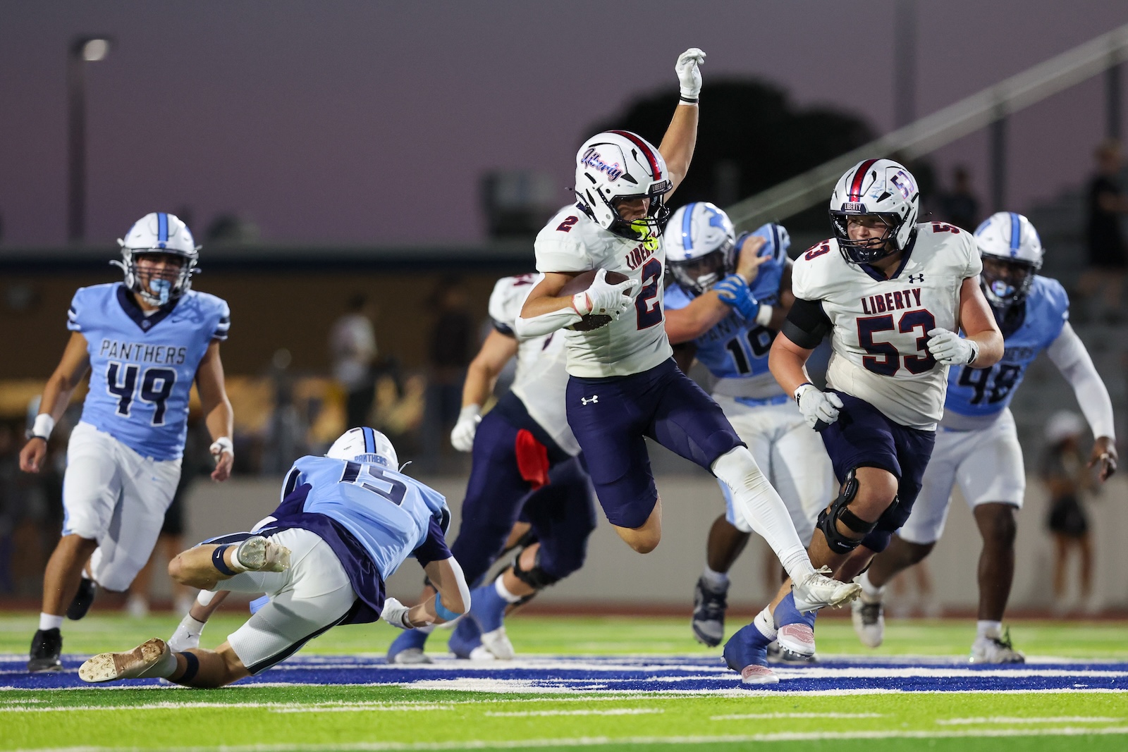 Liberty players in white jerseys chase a ball carrier on a brightly lit football field during a night game with Panthers defenders close behind.
