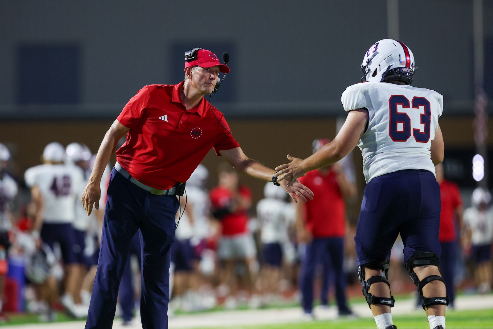 Football coach in red polo and headset shaking hands with a football player wearing number 63 on the sideline during practice or a game.