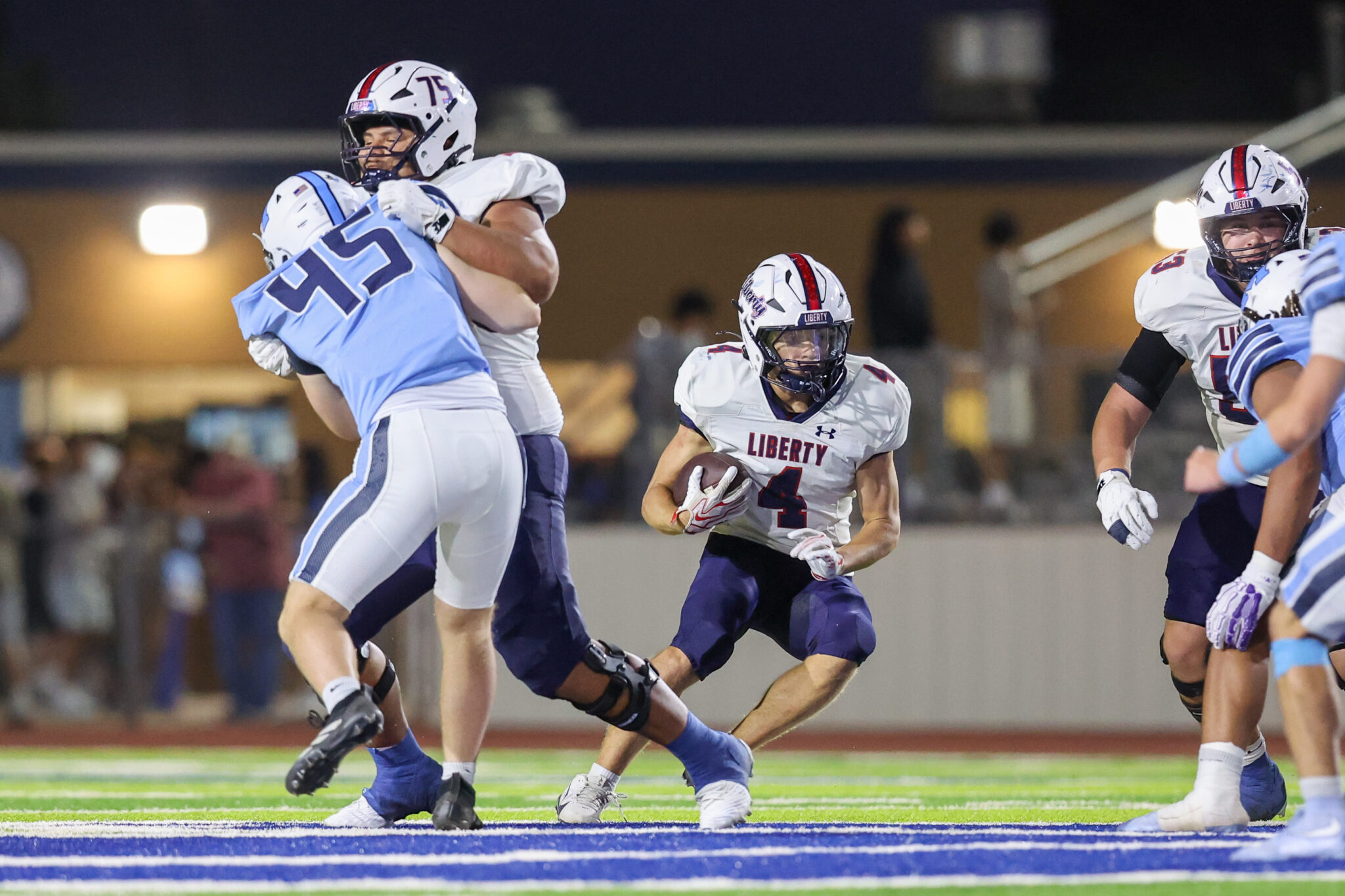 Football game action: a Liberty player in white jersey runs with the ball while a blue-team defender grabs him from behind under stadium lights.