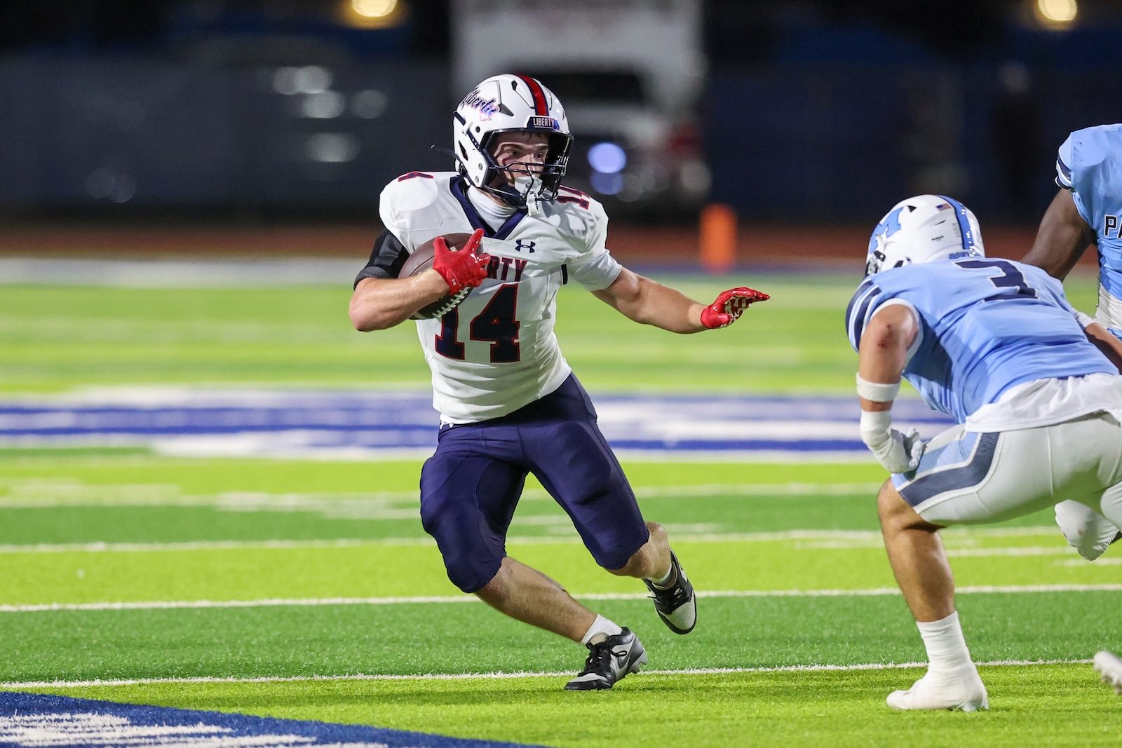 Football player wearing white jersey #14 dashes with the ball while a defender closes in on a lit turf field at night.