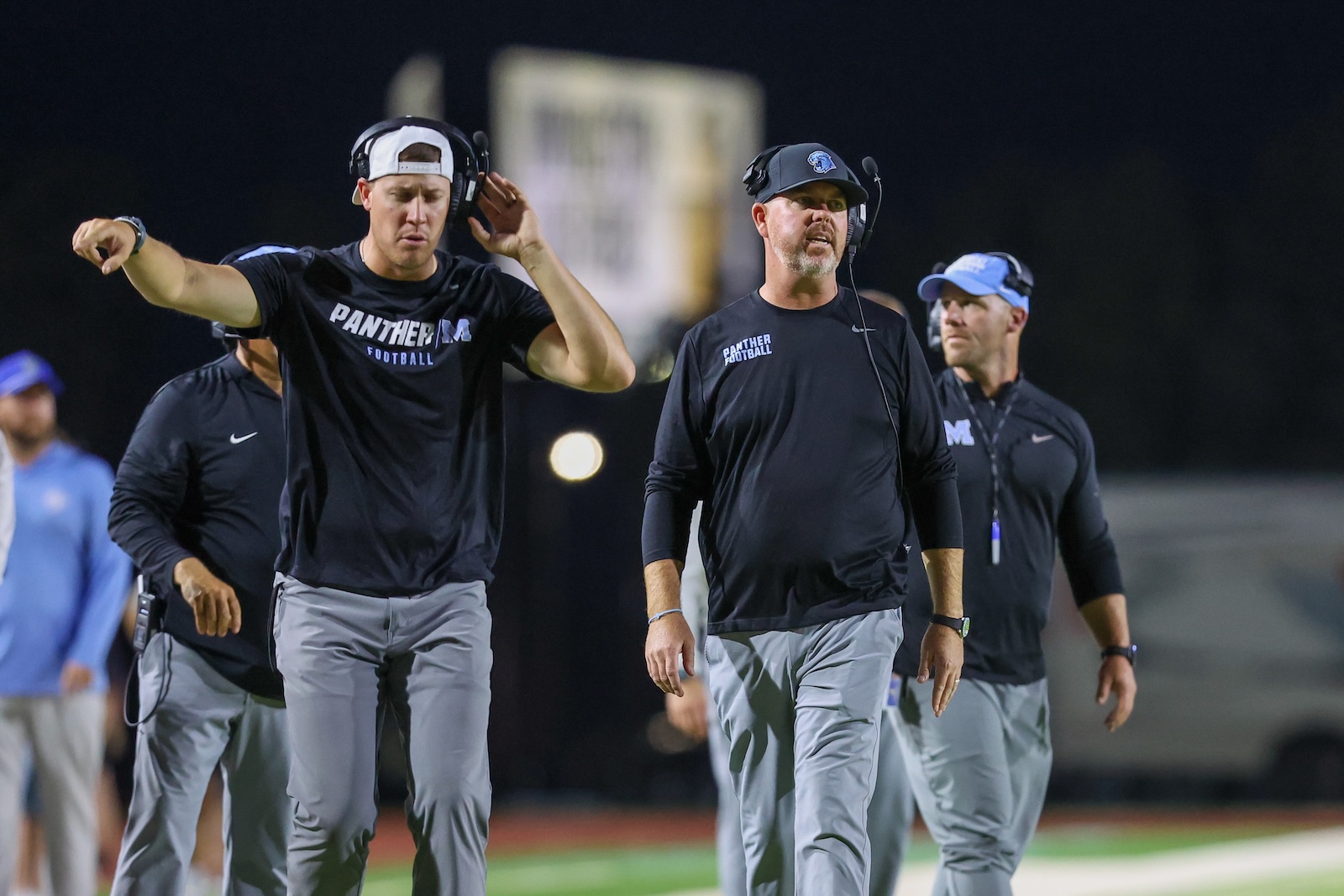 Coaches in black Panthers shirts walking on a football field at night, wearing headsets and listening.