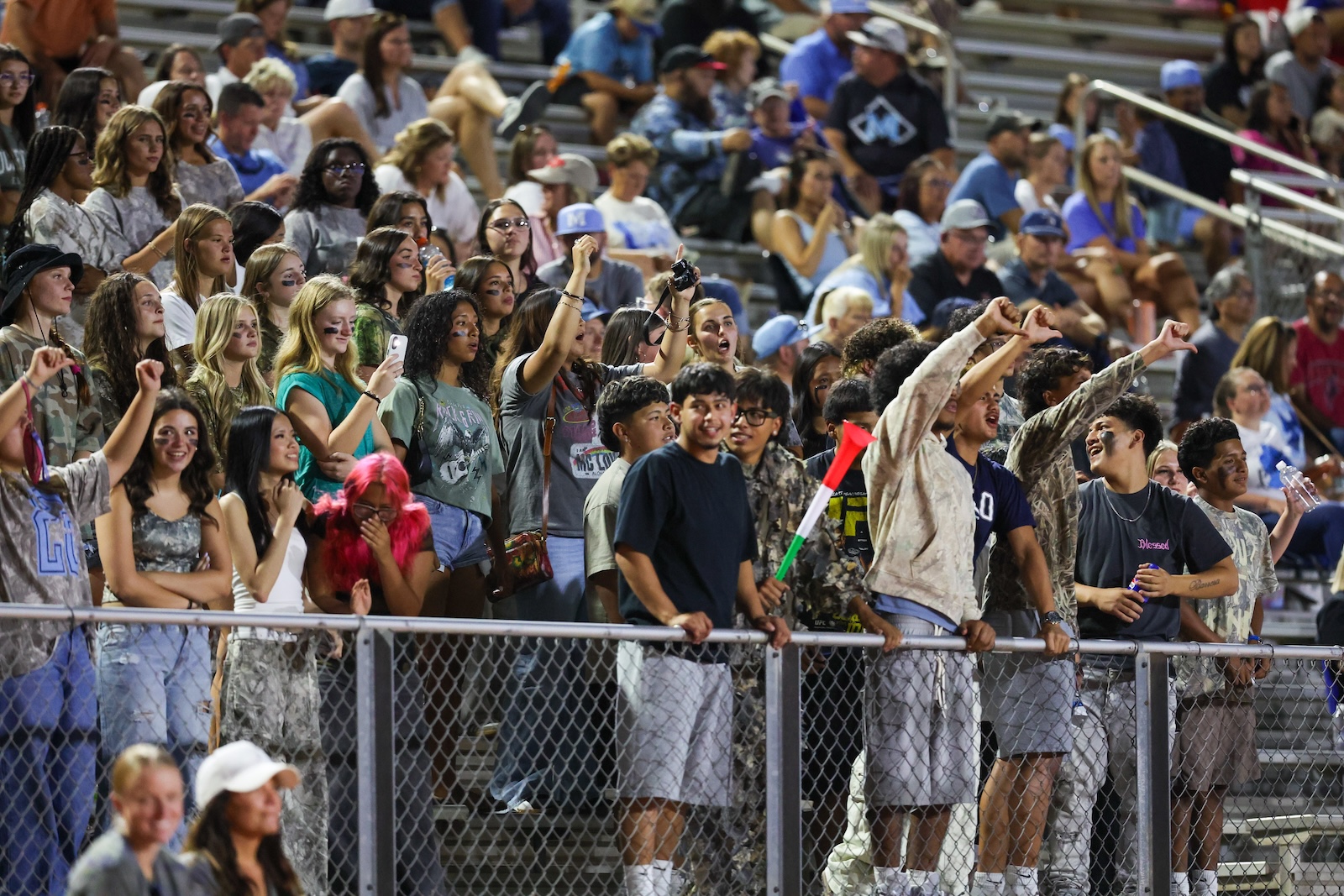 Crowd of teenagers and young adults cheering behind a chain-link fence at a sports game, many raising arms and phones.