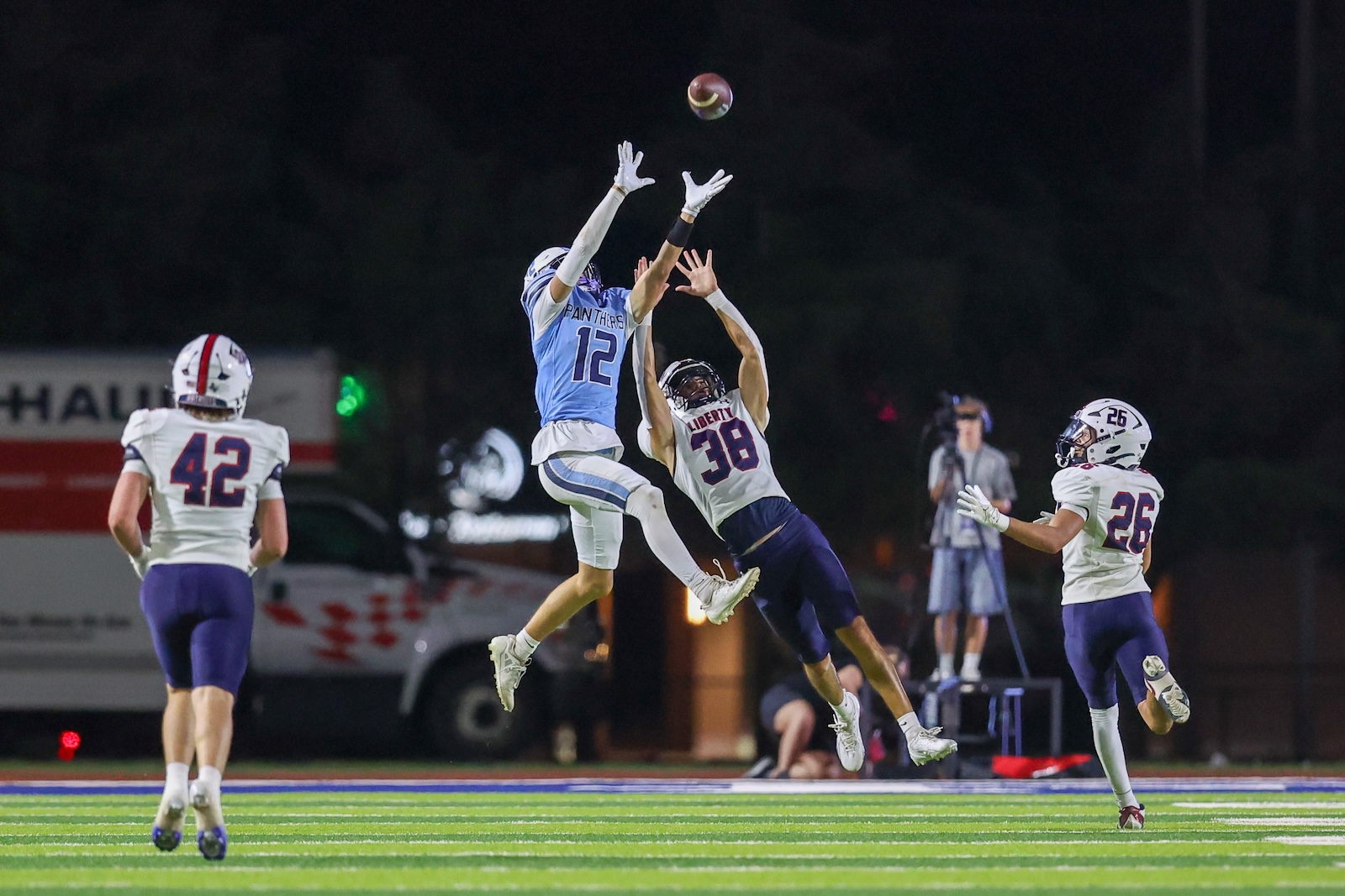 Two football players leap to catch a lofted ball at night, with teammates nearby on a bright green field.