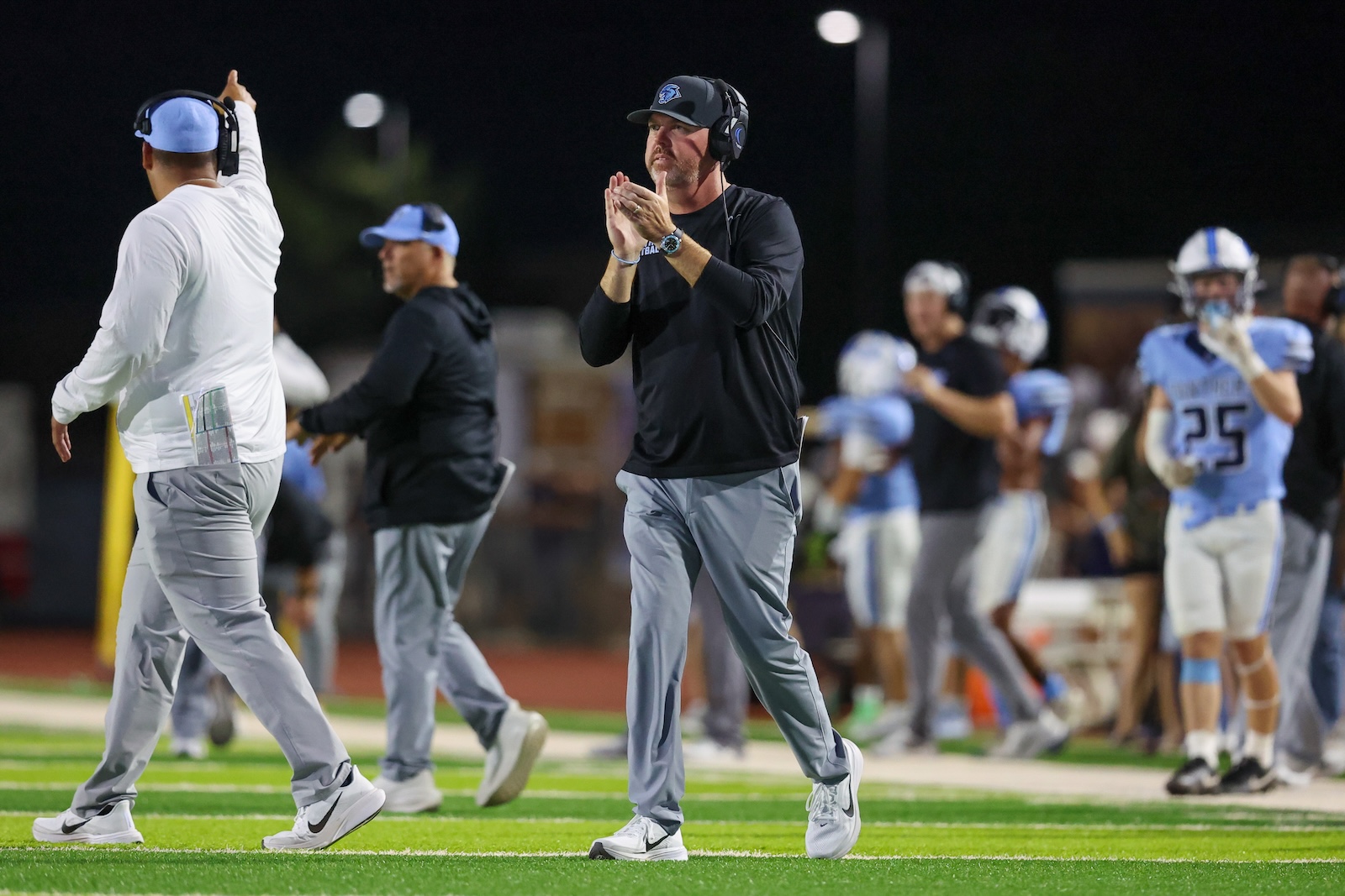 Football coaches and players walk on a night field, with a coach in black shirt clapping as teammates pass by.