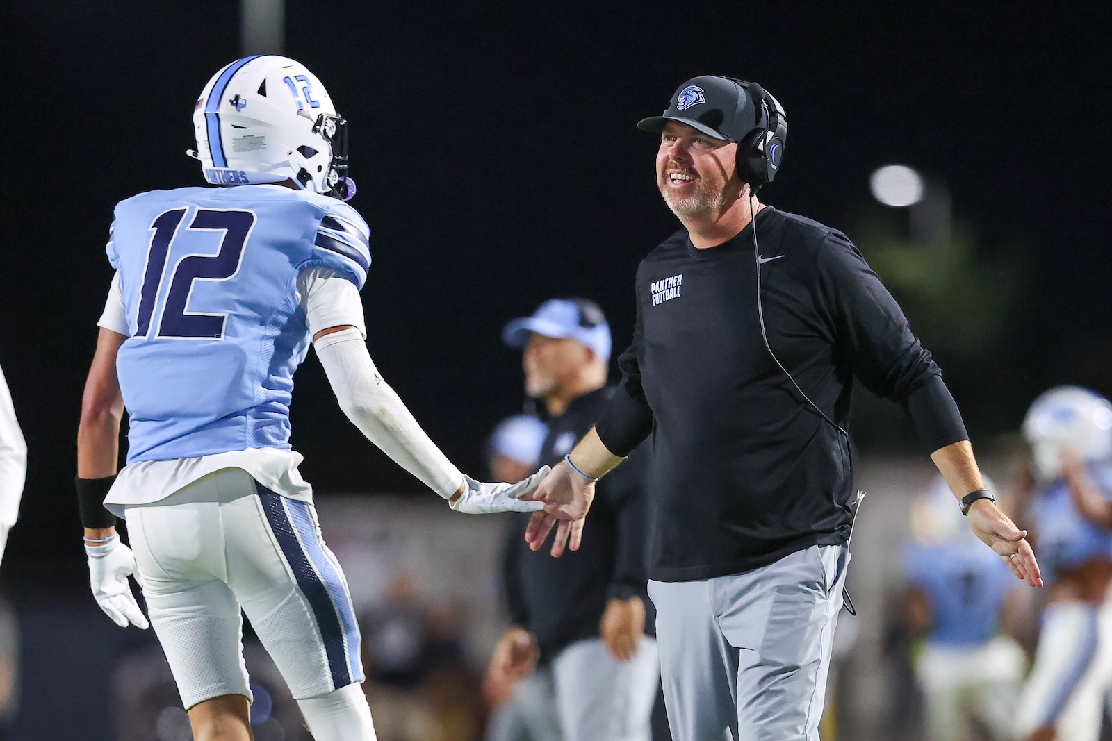 Coach wearing a headset high-fives a football player in a light-blue uniform with the number 12 on a night field.