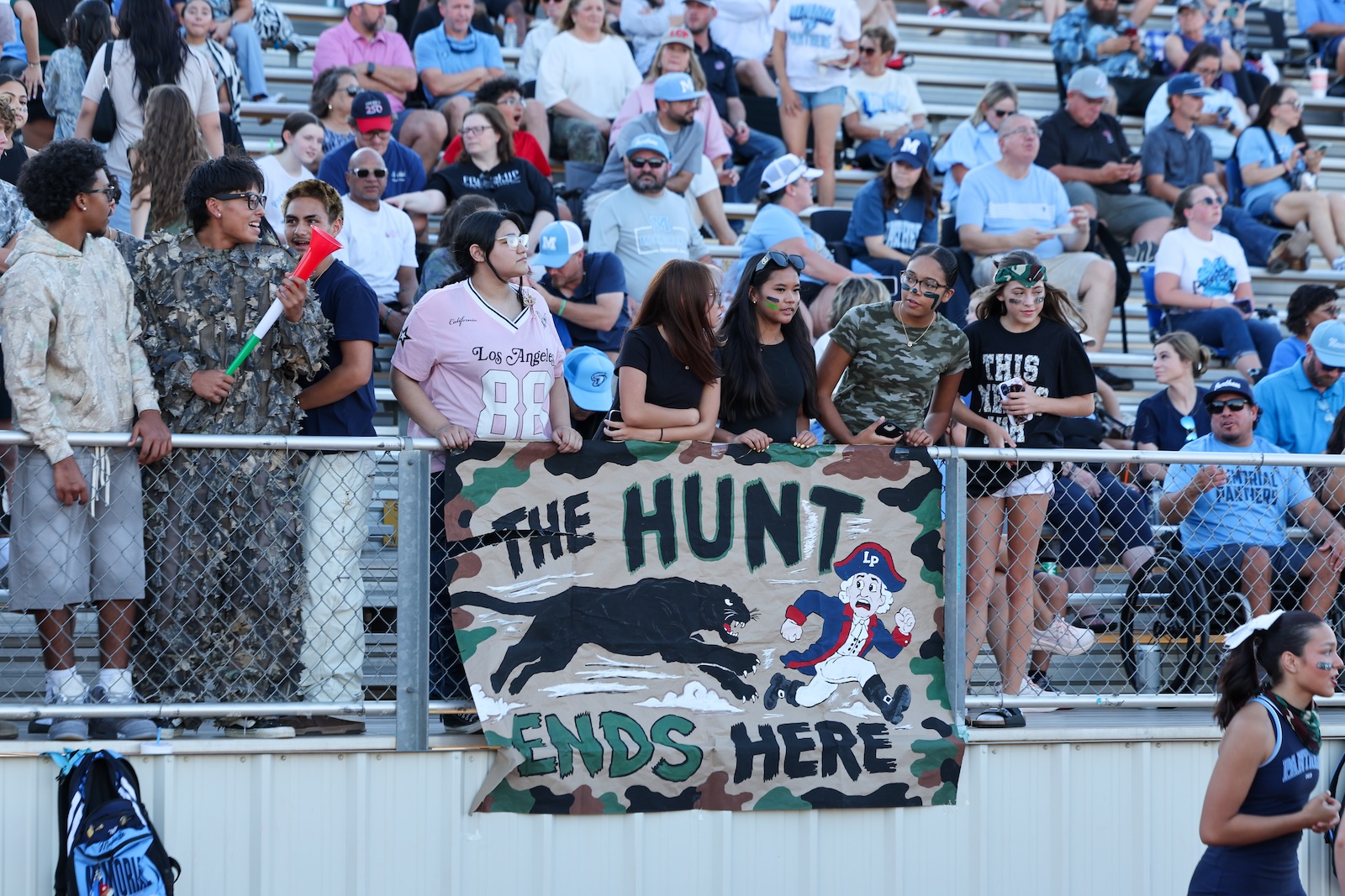 Crowd of spectators at a stadium railing behind a camouflage banner that reads 'THE HUNT ENDS HERE' with a black panther and a cartoon pirate image.